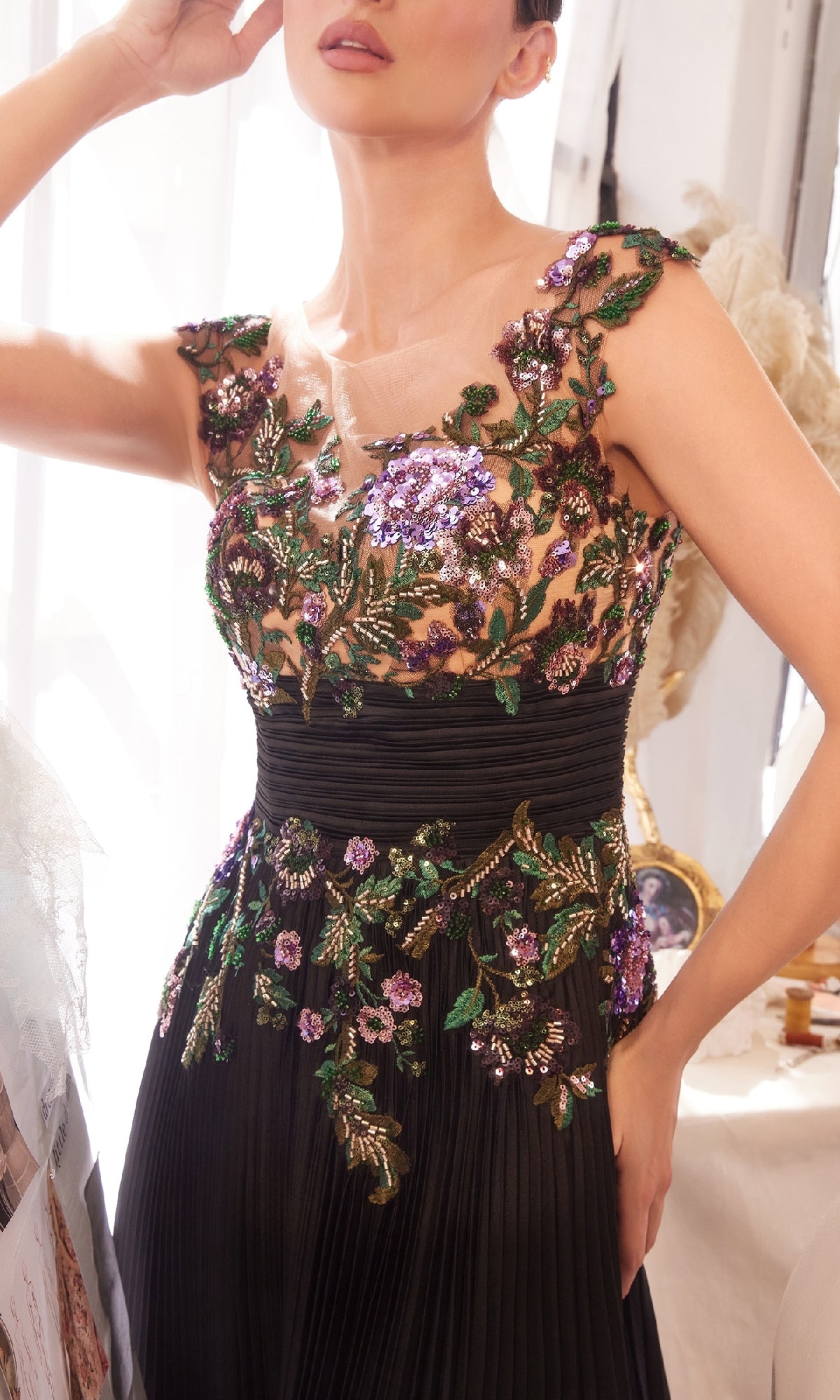 A woman wears the Andrea & Leo A1355 Sequin Long Pleated Prom Dress, a sleeveless black gown with a pleated A-line skirt and green and purple sequin floral appliques on the bodice.